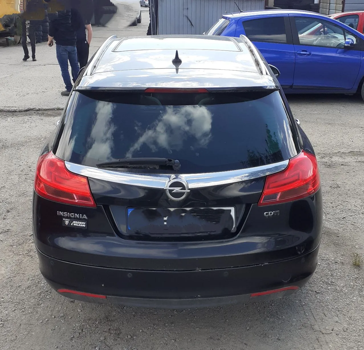 Tylny widok czarnego samochodu marki Opel Insignia. Na klapie bagażnika widoczne jest logo Opela, napis 'INSIGNIA' po lewej stronie oraz 'CDTI' po prawej. Samochód posiada panoramiczny dach i jest zaparkowany na żwirowym placu. W tle widoczny jest niebieski samochód oraz dwie osoby.