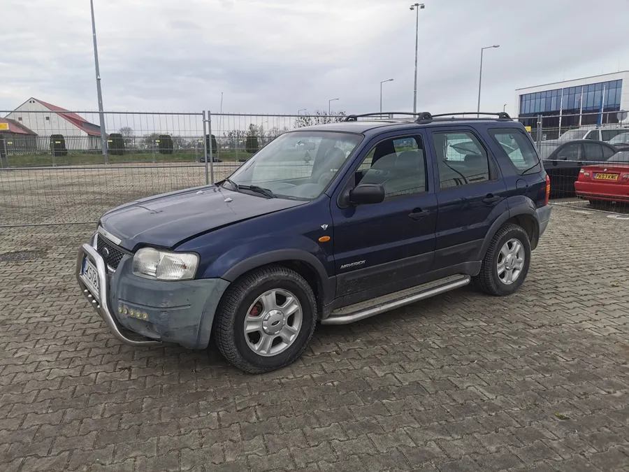 Ciemnoniebieski samochód typu SUV zaparkowany na brukowanej powierzchni. Pojazd posiada przedni zderzak z metalową osłoną, dachowy bagażnik i srebrne podnóżki. W tle widoczne są ogrodzenie, budynki, latarnie uliczne oraz czerwony samochód. Niebo jest zachmurzone.