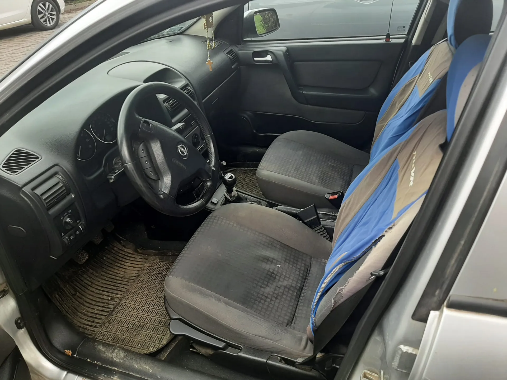 Wnętrze samochodu z otwartą drzwiami kierowcy. Widoczna czarna kierownica z logo, deska rozdzielcza z licznikami, czarne siedzenia z niebiesko-brązowymi pokrowcami, brudne dywaniki podłogowe oraz dźwignia zmiany biegów. Na lusterku wstecznym wiszą różaniec i krzyż. Poza samochodem widoczna część innego pojazdu i brukowana powierzchnia.