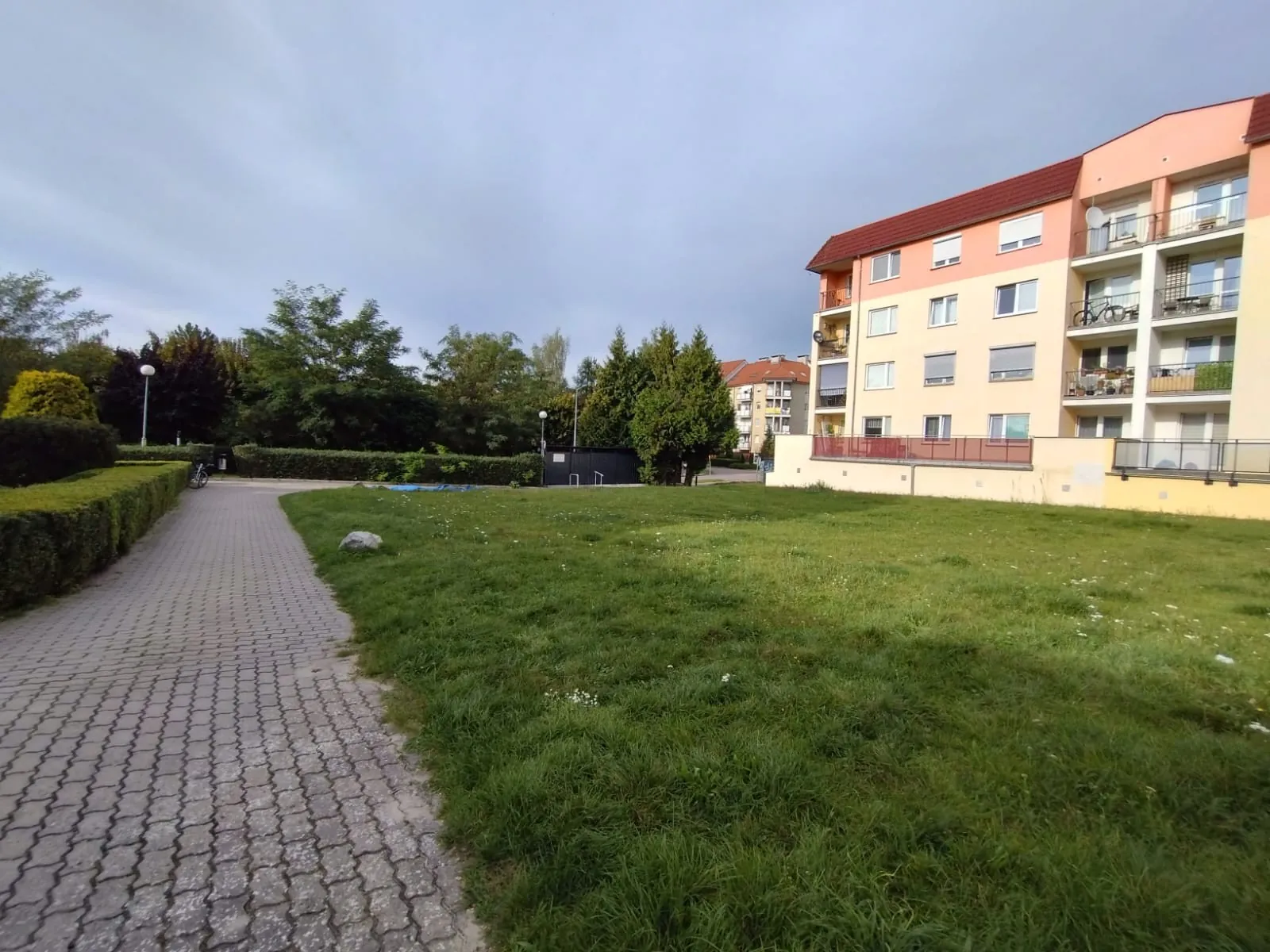 Widok na wielorodzinny budynek mieszkalny o żółtej elewacji i czerwonym dachu, z balkonami po prawej stronie. Przed budynkiem znajduje się rozległy trawnik, a po lewej stronie biegnie ścieżka wyłożona szarą kostką brukową, oddzielona od trawy niskim, przystrzyżonym żywopłotem. W tle widoczne są liczne zielone drzewa i krzewy pod jasnym, lekko zachmurzonym niebem.