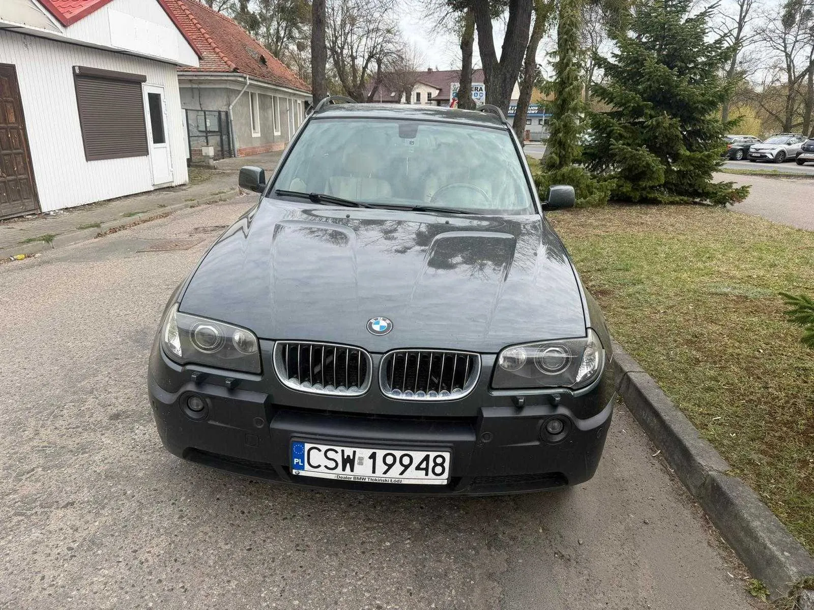 Ciemnoszary samochód BMW X3 zaparkowany na asfaltowej nawierzchni. Widoczna tablica rejestracyjna z polskim kodem PL i numerem CSW 19948. W tle białe budynki z czerwonymi dachami, drzewa, trawnik oraz inne zaparkowane pojazdy. Samochód ma charakterystyczne podwójne klapki chłodnicy i logo BMW na masce.