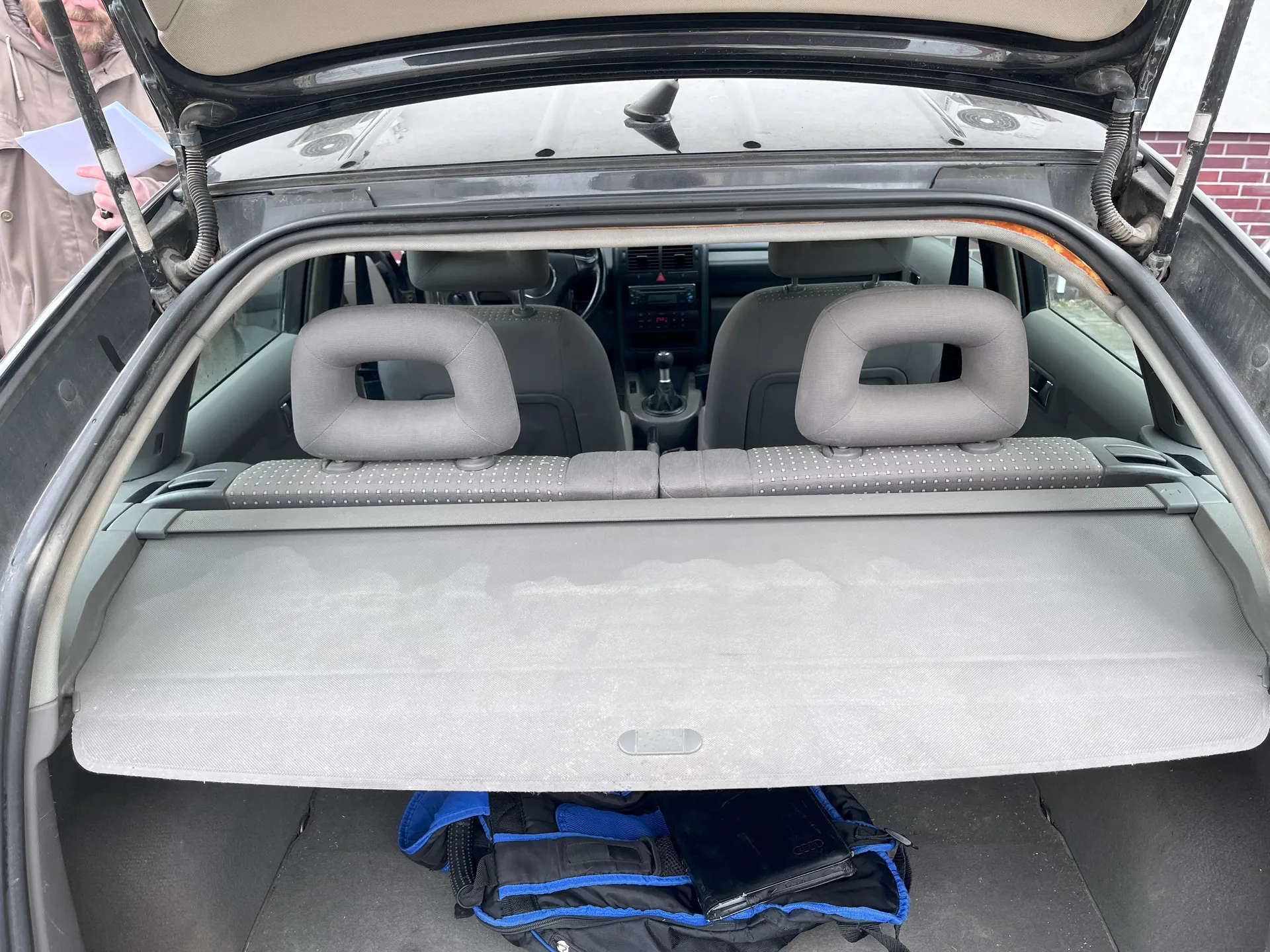Widok na wnętrze samochodu z otwartym bagażnikiem. W tle widoczne są dwa przednie siedzenia z podłokietnikami, deska rozdzielcza z radiem i dźwignią zmiany biegów. Na środku bagażnika znajduje się szara osłona przeciwsłoneczna, pod którą leży czarno-niebieski plecak z czarnym tabletem. Po lewej stronie obrazu osoba w beżowym płaszczu trzyma białą kartkę papieru. W tle po prawej stronie widoczna jest ceglana ściana.
