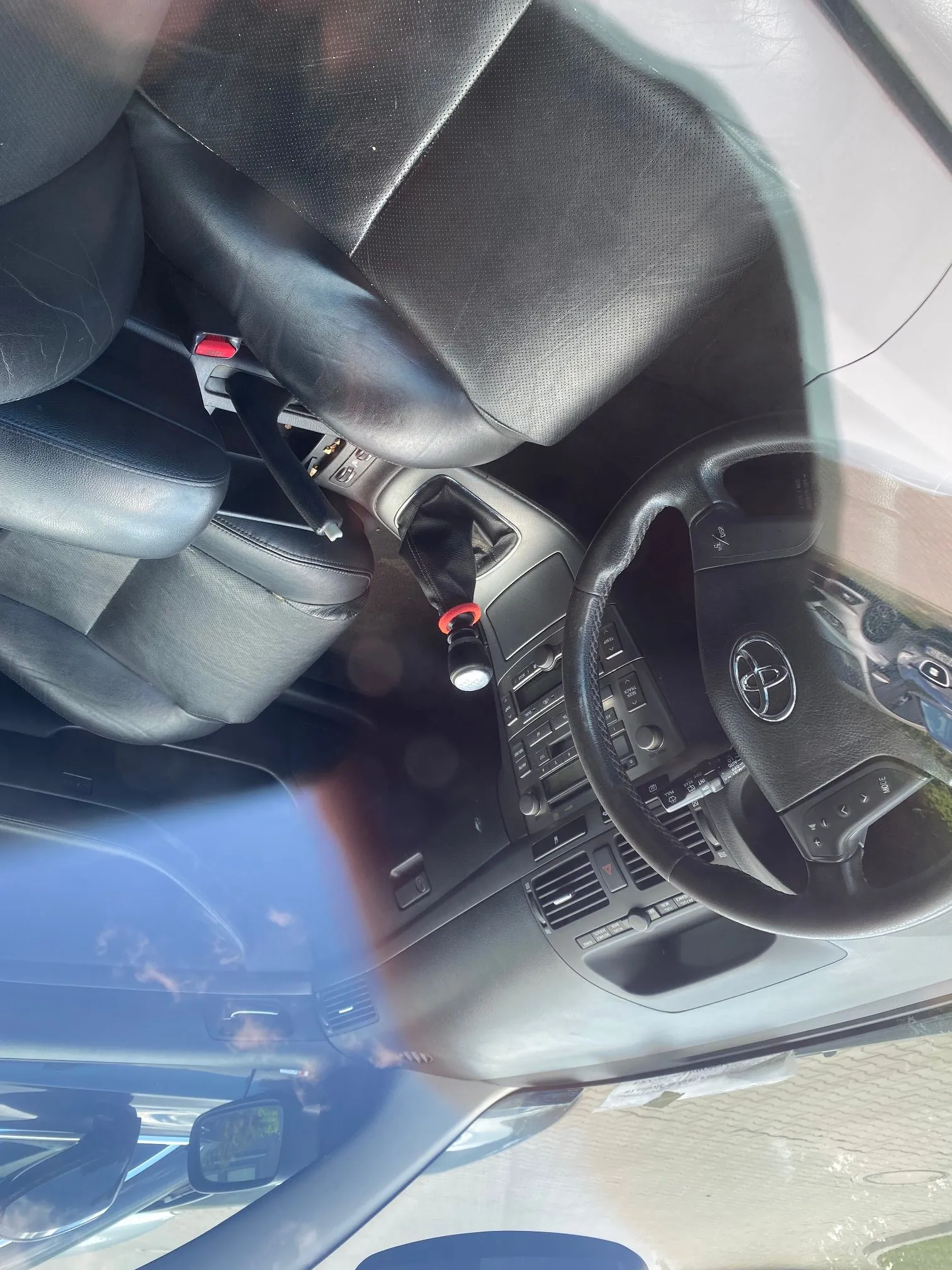 Wnętrze samochodu z czarną skórzaną tapicerką. Widoczna jest kierownica z logo Toyoty, konsola środkowa z lewarkiem zmiany biegów z czerwoną nakładką oraz przednie fotele. Zdjęcie jest wykonane pod kątem, z widocznym odblaskiem na przedniej szybie.