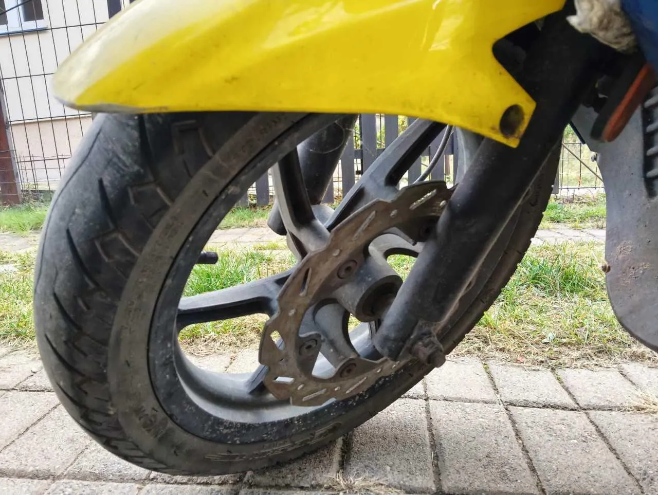 Zbliżenie na przednie koło motocykla z żółtym błotnikiem. Widoczna jest czarna opona, czarna felga oraz tarcza hamulcowa z widocznymi otworami wentylacyjnymi. Motocykl stoi na kostce brukowej, w tle widać trawę i metalowy płot.