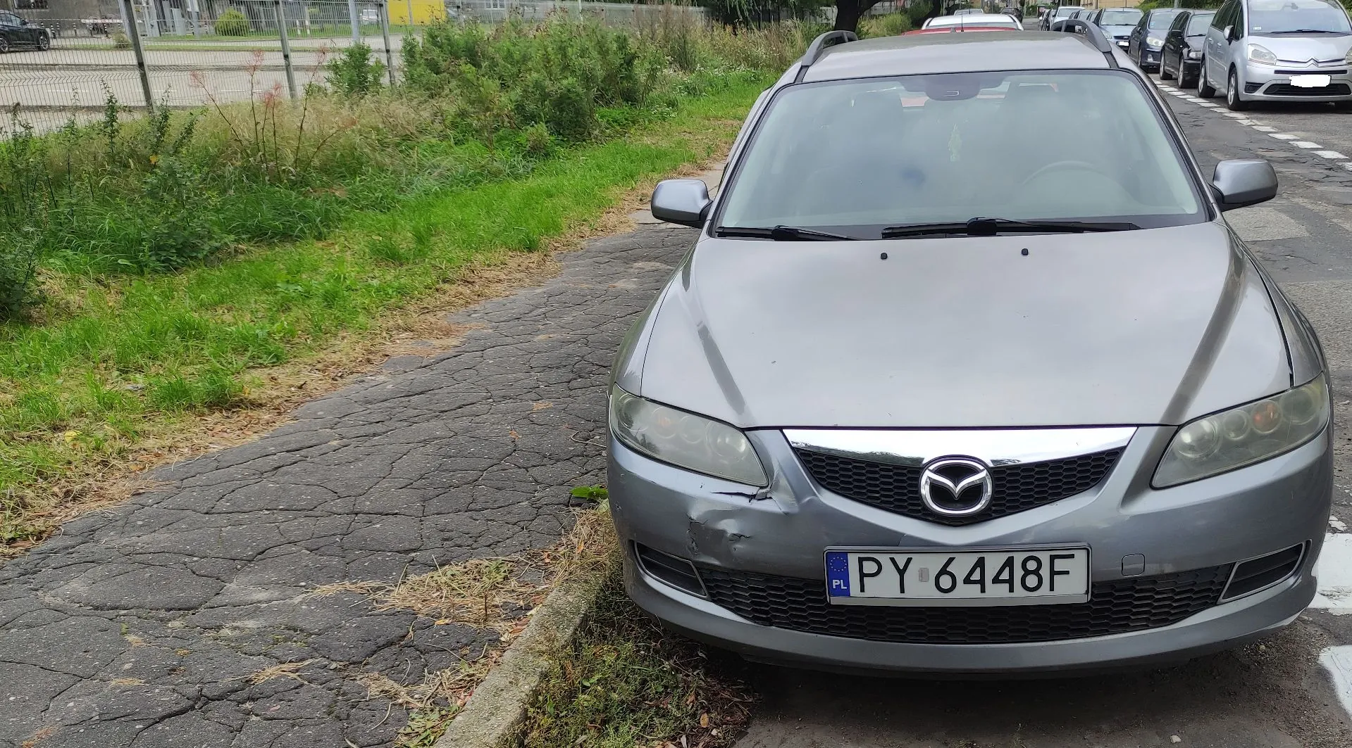 Srebrny samochód marki Mazda zaparkowany przodem do obiektywu na krawędzi zniszczonego chodnika. Pojazd posiada polską tablicę rejestracyjną o numerze PY 6448F. Na przednim zderzaku, po lewej stronie, widoczne jest wgniecenie. W tle znajduje się zielony pas trawy, ogrodzenie oraz inne zaparkowane samochody.
