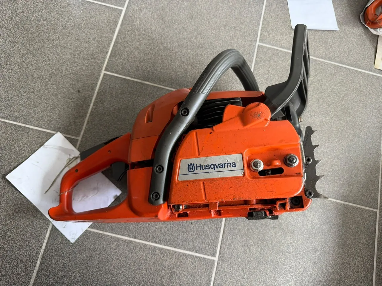 Pomarańczowa piła spalinowa marki Husqvarna leżąca na szarych płytkach ceramicznych. Na obudowie urządzenia widnieje napis 'Husqvarna' wraz z logo. Obok piły znajduje się biała kartka papieru.