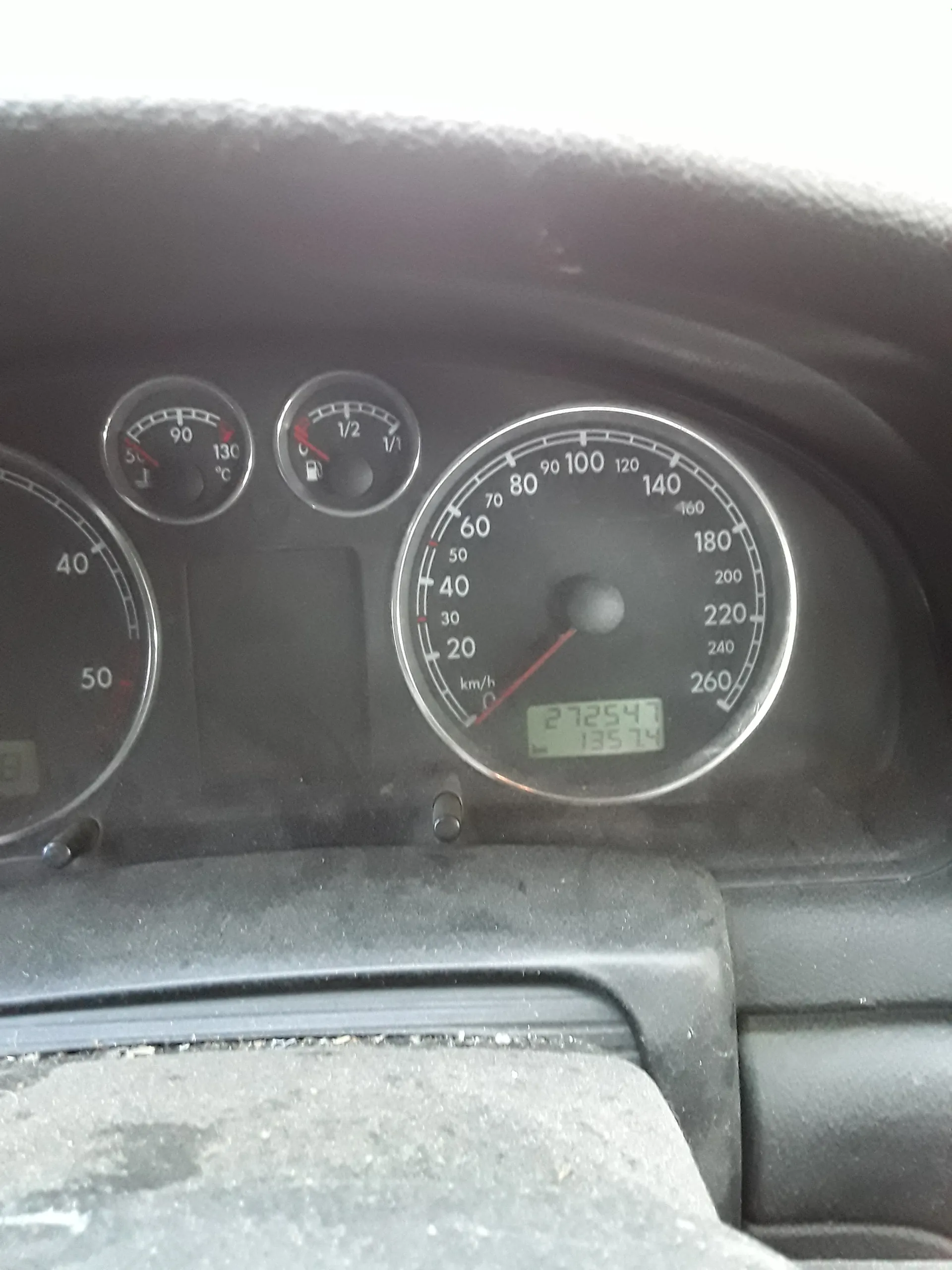 Zbliżenie na deski rozdzielcze samochodu. W centrum znajduje się prędkościomierz z zakresem do 260 km/h, wskazówka na 0 km/h. Obok cyfrowy wyświetlacz z liczbami 272541 i 135.7. Poniżej mniejsze wskaźniki: poziom paliwa (1/2), temperatura chłodniczy (około 90°C) oraz inny wskaźnik z zakresem do 50. Całość umieszczona na czarnym tle deski rozdzielczej.