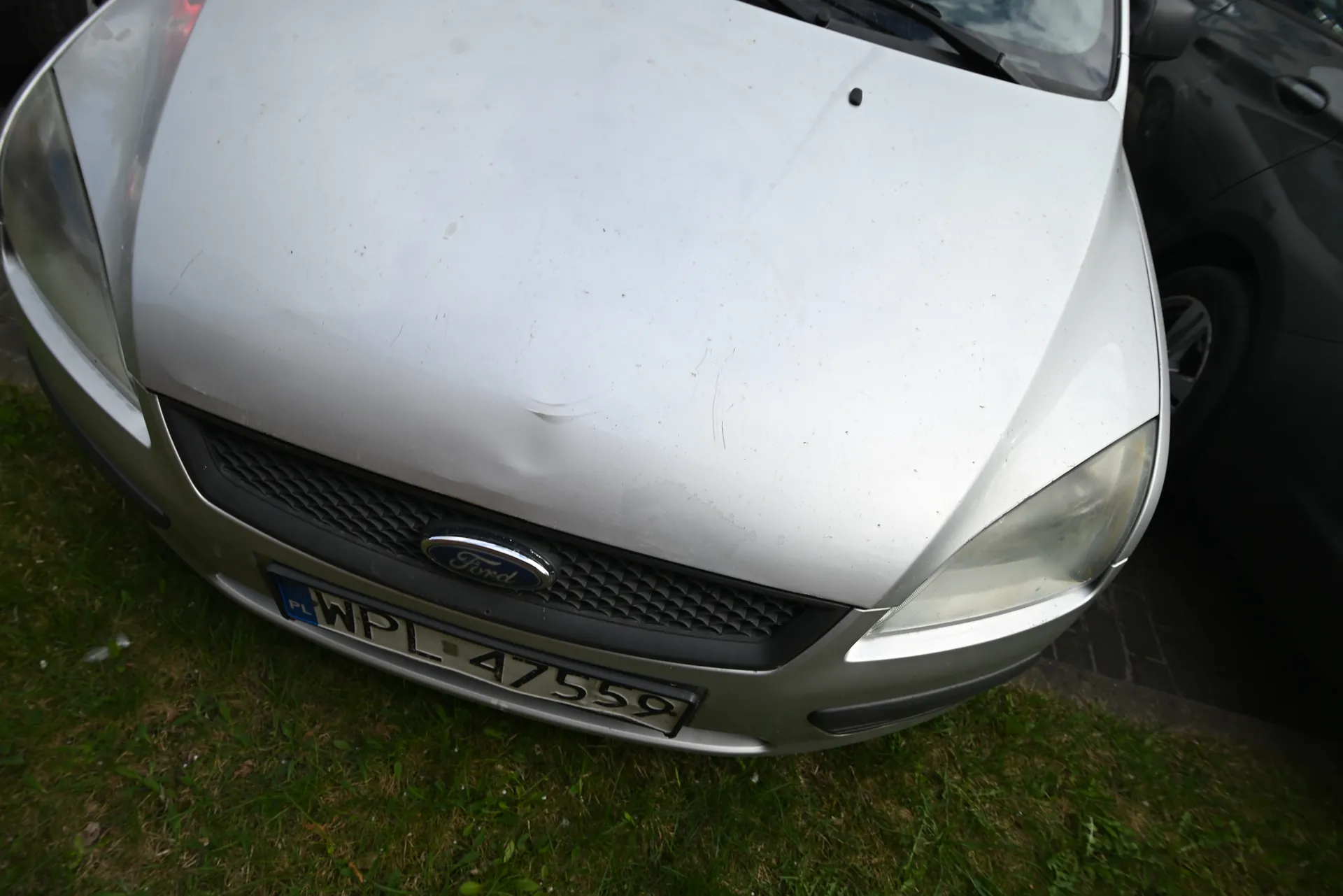 Przód srebrnego samochodu marki Ford z widocznym logo na atrapie chłodnicy. Tablica rejestracyjna z numerem WPL 47559 oraz oznaczeniem PL po lewej stronie. Na masce widoczne są zadrapania i wgniecenie. Samochód zaparkowany na trawie, obok częściowo widoczny czarny pojazd.