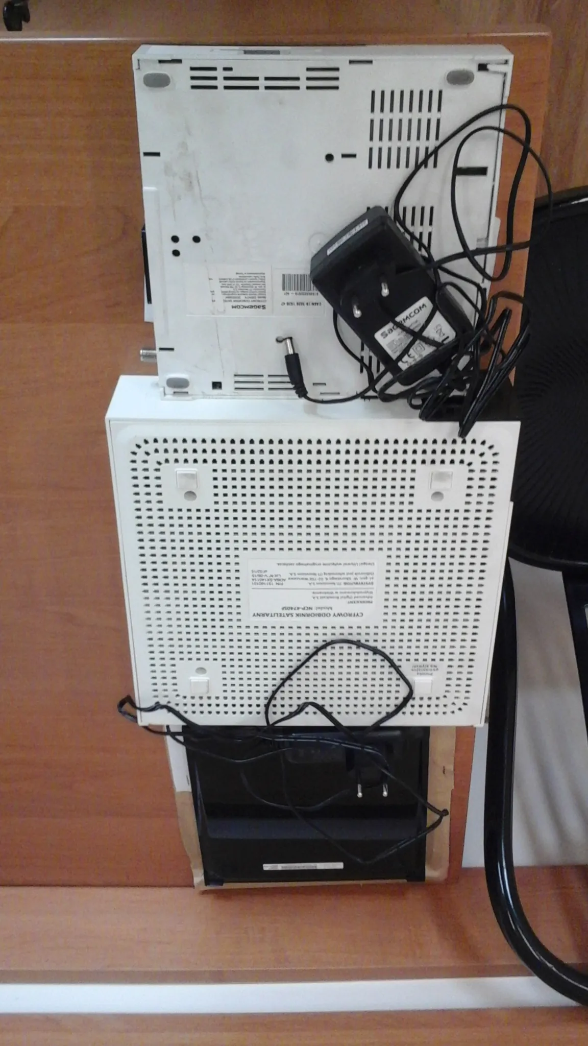 Dwa białe urządzenia elektroniczne z siatką wentylacyjną, na których widoczne są etykiety z tekstem, w tym 'CYFROWY ODBIORNIK SATELITARNY'. Obok znajduje się czarny zasilacz z kablem i wtyczką. Urządzenia umieszczone są na pionowej powierzchni drewnianej. W tle widoczne są czarne przewody i czarna konstrukcja.