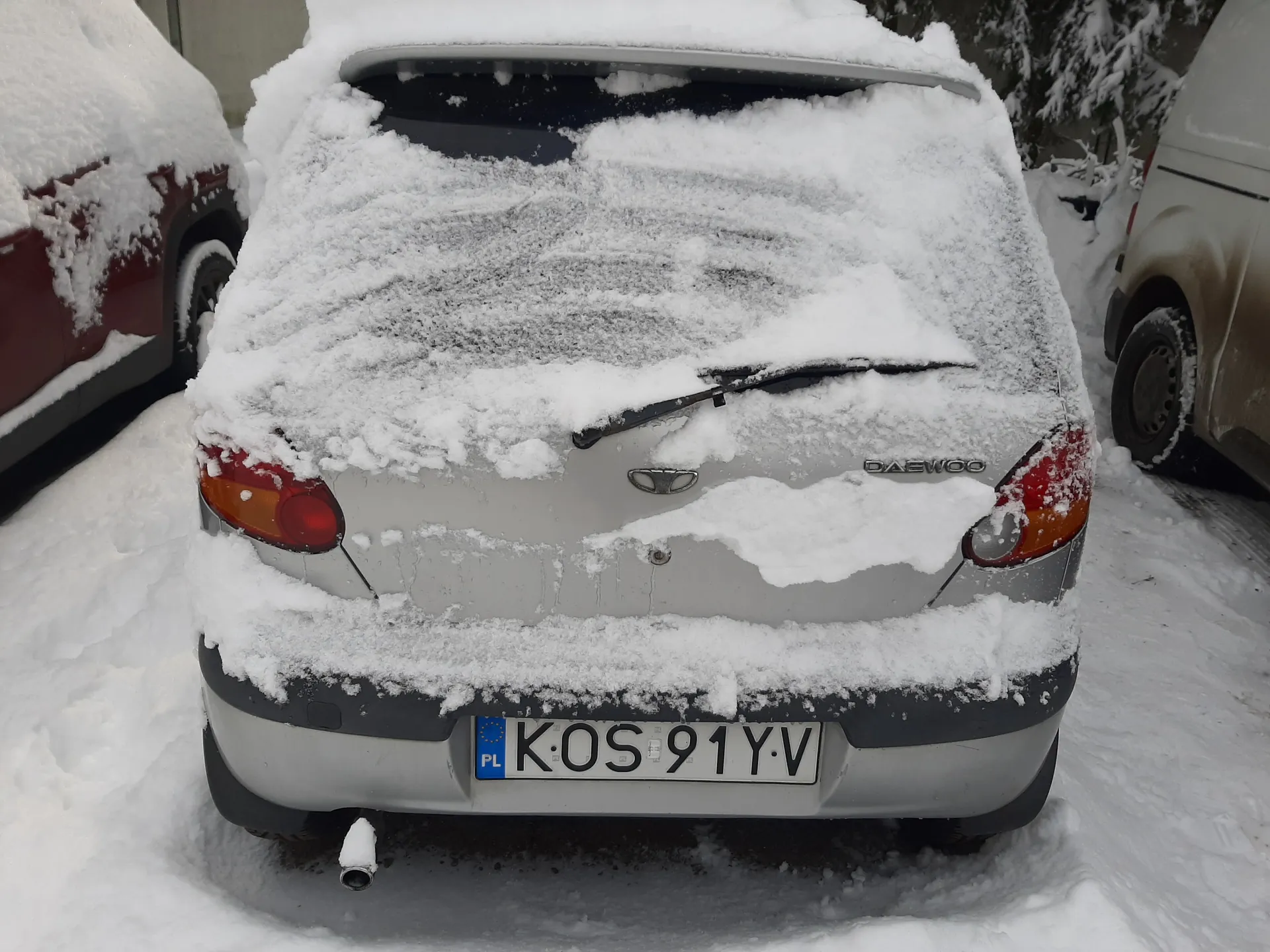 Tył srebrnego samochodu marki Daewoo pokrytego śniegiem. Na bagażniku widoczny napis "DAEWOO" oraz logo marki. Tablica rejestracyjna z polskim kodem "PL" i numerem "K-OS 91YV". Samochód otoczony śniegiem, z widocznymi fragmentami innych pojazdów po obu stronach: czerwonego po lewej i beżowego po prawej. Tło stanowią śnieżne drzewa i zasneżona powierzchnia ziemi. Wycieraczka na tylnej szybie jest podniesiona.