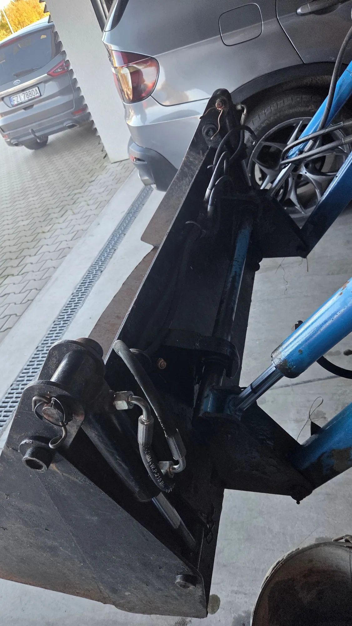 Zbliżenie na czarną, metalową konstrukcję maszyny rolniczej lub budowlanej z niebieskimi siłownikami hydraulicznymi. Urządzenie znajduje się na betonowym podłożu obok zaparkowanego srebrnego samochodu. W tle widoczny jest fragment drugiego, szarego samochodu z tablicą rejestracyjną FZI 7880A.