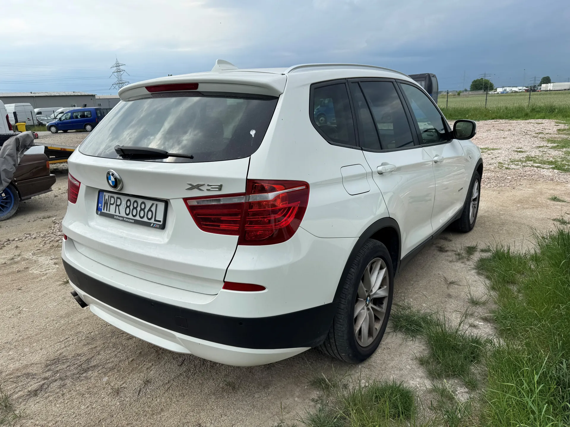 Biały samochód SUV marki BMW model X3 zaparkowany na żwirowym terenie z zielonymi łatkami trawy. Widoczny tył pojazdu z tablicą rejestracyjną WPR 8886L oraz logo BMW. W tle niebieski samochód dostawczy, inne pojazdy, budynki, linie energetyczne i częściowo zachmurzone niebo.