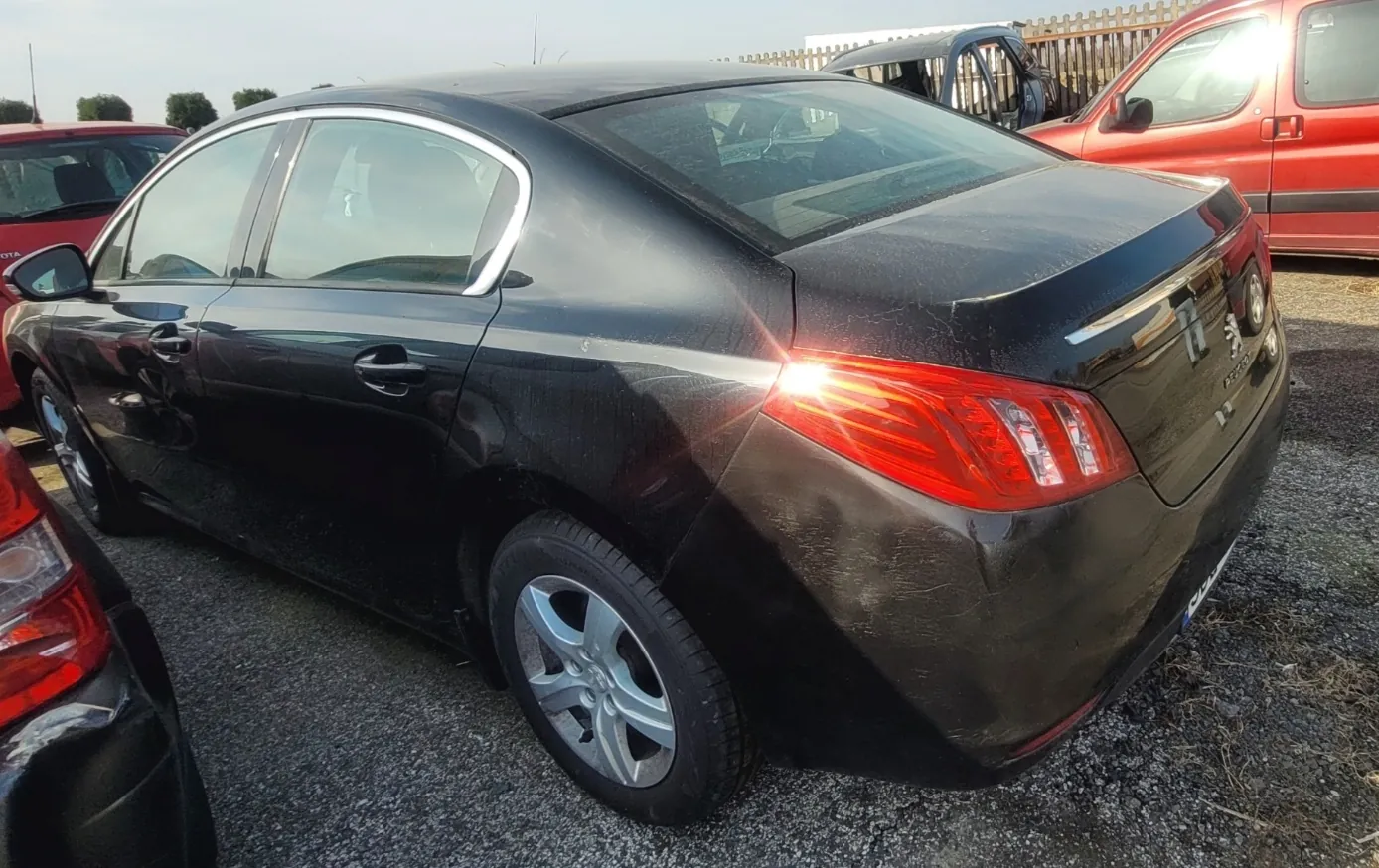 Czarny samochód osobowy marki Peugeot, widok z tyłu i boku. Pojazd stoi na żwirowym podłożu. W tle widoczne są inne samochody: czerwony po lewej stronie i czerwony z czarną listwą po prawej. Za nimi drewniane ogrodzenie i drzewa. Na tylnej szybie i bagażniku widoczne są odbicia światła. Na bagażniku umieszczony jest napis 'Peugeot' oraz logo marki. Niebo jest jasne, bez chmur.