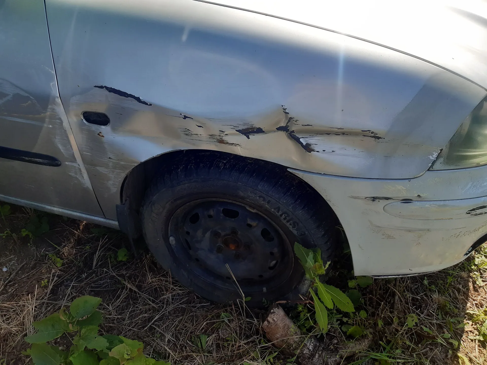 Zbliżenie na przedni prawy błotnik srebrnego samochodu, który jest mocno wgnieciony i porysowany nad kołem. Opona jest całkowicie pozbawiona powietrza i spłaszczona. Samochód stoi na trawiastym podłożu.