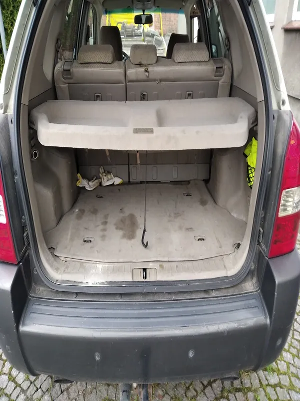 Widok na otwarty bagażnik samochodu typu SUV. Wnętrze bagażnika jest beżowe, z widoczną półką na środku. W lewej części bagażnika leżą białe rękawice lub tkanina. Na podłodze bagażnika znajdują się plamy. Widoczne są tylna część siedzeń, czerwone światła tylnego oraz ciemnoszary zderzak. Samochód stoi na brukowanej powierzchni, w tle widać fragment budynku i drzewa.