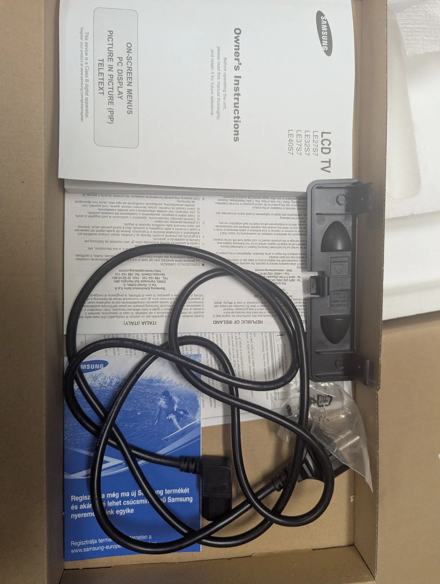 Zdjęcie z góry wnętrza kartonowego pudełka, w którym znajduje się czarny kabel zasilający z wtyczką, instrukcja obsługi telewizora Samsung LCD TV oraz inne dokumenty papierowe. Na instrukcji widoczne są napisy: 'Owner's Instructions', 'LCD TV', 'LE26S7', 'LE32S7', 'LE37S7', 'LE40S7' oraz 'ON-SCREEN MENUS PC DISPLAY PICTURE IN PICTURE (PIP) TELETEXT'.