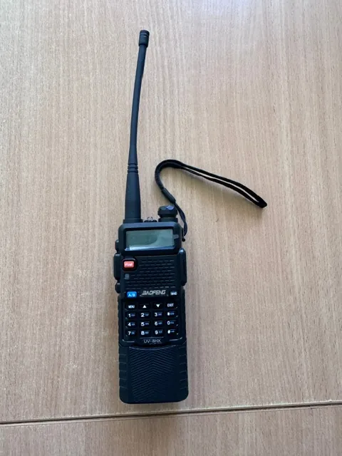 Czarny radiotelefon marki Baofeng model UV-5HX z długą anteną, leżący na jasnobrązowej, drewnianej powierzchni. Urządzenie posiada mały ekran LCD, klawiaturę numeryczną, czerwony przycisk PTT oraz czarny sznurek do nadgarstka przymocowany do obudowy.