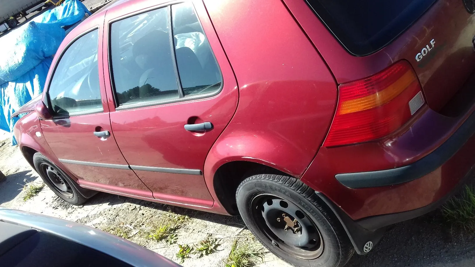 Zdjęcie przedstawiające tylną i boczną część czerwonego samochodu marki Volkswagen Golf. Widoczny jest napis GOLF na klapie bagażnika, tylna lampa oraz dwa boczne drzwi z czarnymi listwami ochronnymi i klamkami. Samochód stoi na piaszczystym podłożu.