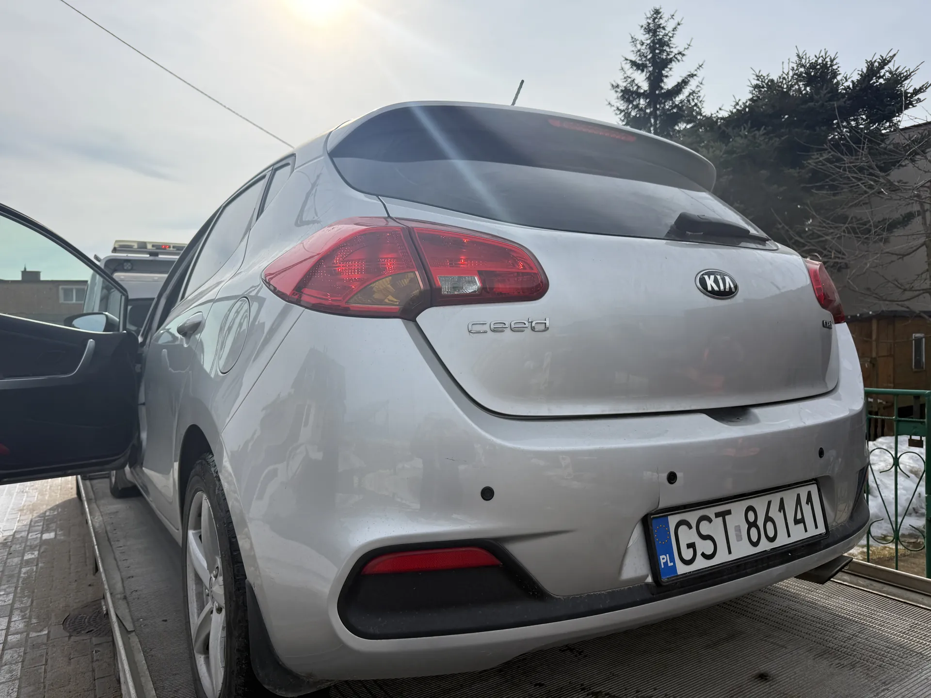 Tył srebrnego samochodu Kia Ceed z otwartą lewą przednią drzwiami. Widoczny jest logo KIA, napis ceed oraz tablica rejestracyjna PL GST 86141. Samochód stoi na metalowej powierzchni, w tle drzewa, budynek, fragment zielonego ogrodzenia i śnieg. Na niebie słońce i chmury, za samochodem częściowo widoczny inny pojazd z dachowym bagażnikiem.