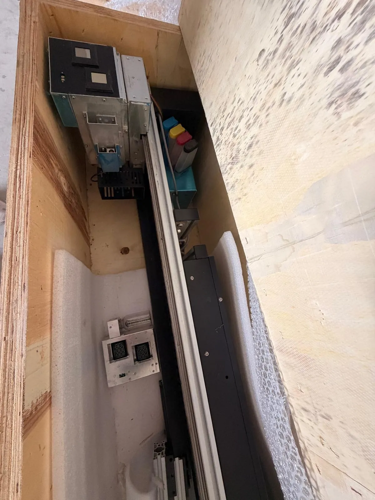 Zdjęcie przedstawiające wnętrze drewnianej obudowy z zamontowanymi komponentami elektronicznymi i mechanicznymi. Widoczny jest czarny moduł sterujący z gniazdami, metalowe szyny prowadzące oraz elementy izolacyjne z białej pianki. W głębi znajdują się kolorowe elementy w odcieniach niebieskiego, żółtego i czerwonego.