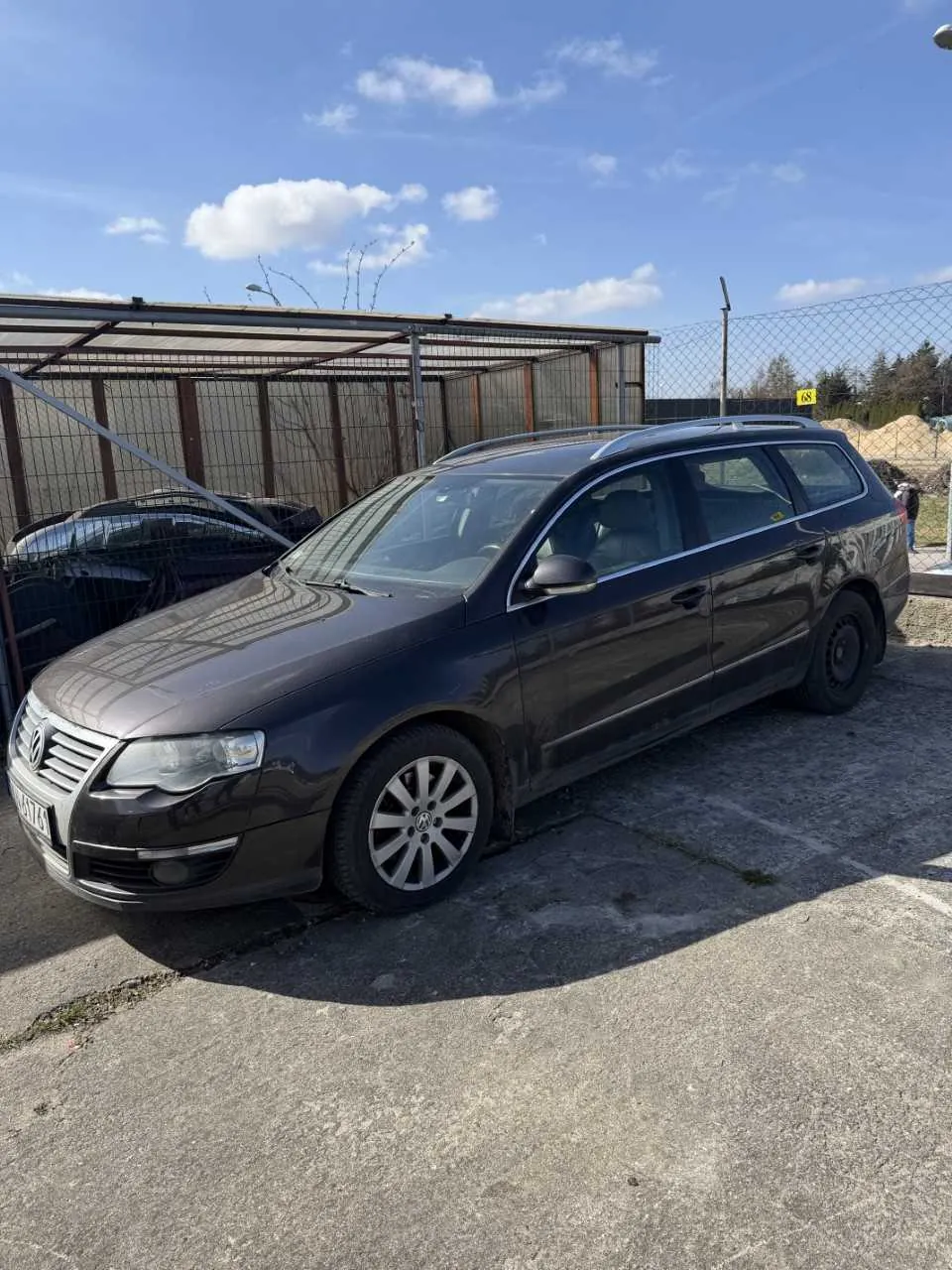 Ciemnoszary samochód osobowy typu kombi marki Volkswagen zaparkowany na betonowej powierzchni. W tle widoczny jest czarny samochód pod metalowym daszkiem z siatką, ogrodzenie z drutu kolczastego, żółty znak z cyfrą 68 oraz niebo z kilkoma chmurami. Na pierwszym planie betonowa nawierzchnia z widocznymi pęknięciami.