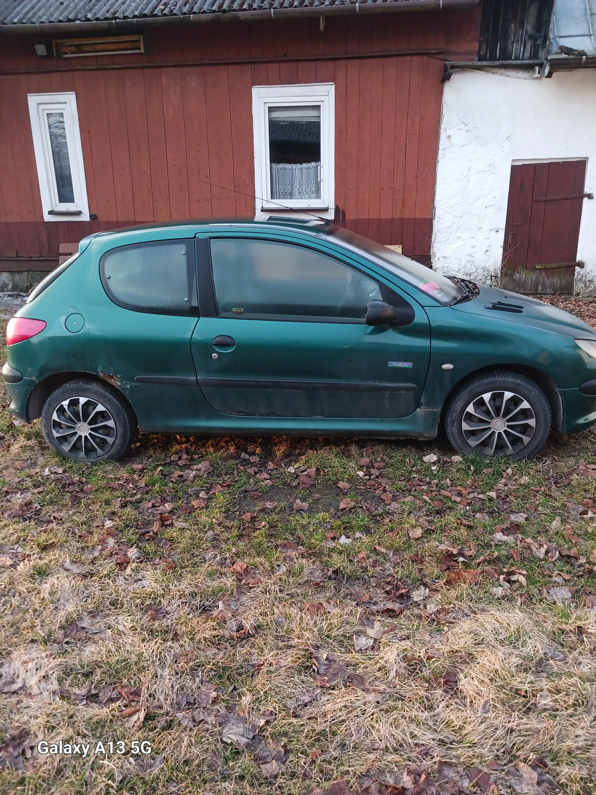 Zielony samochód osobowy zaparkowany na trawie obok budynku o czerwonych drewnianych ścianach i białych obramowaniach okien. W budynku widoczne są okna z białymi ramami, jedno z nich zasłonięte koronką oraz brązowe drzwi. Trawa na podwórku częściowo wyschnięta, z suchymi liśćmi. W prawym dolnym rogu zdjęcia znajduje się napis 'Galaxy A13 5G'.