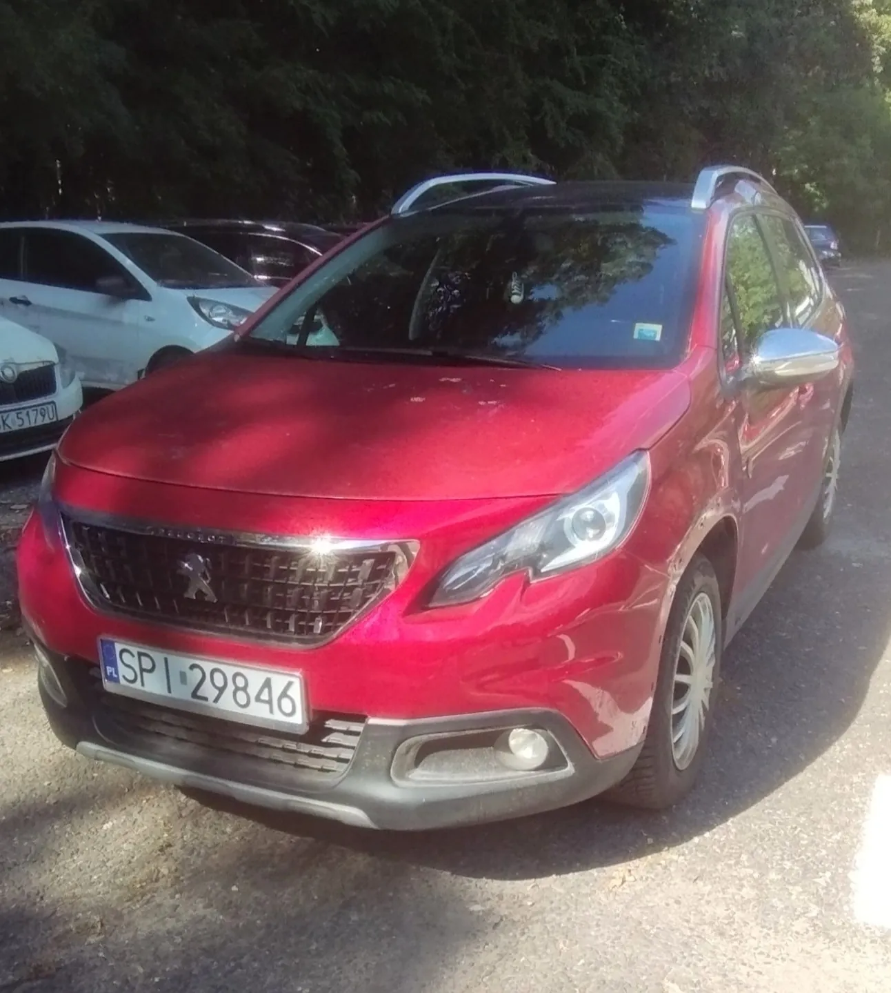 Czerwony samochód marki Peugeot zaparkowany na zewnątrz. Widoczna jest przednia część pojazdu z tablicą rejestracyjną o numerze SPI 29846. W tle znajdują się inne samochody oraz gęste, zielone drzewa.