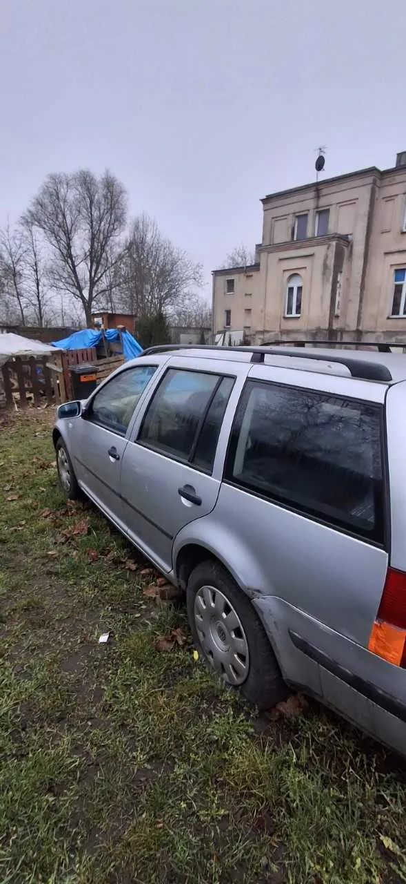 Srebrny samochód typu kombi zaparkowany na trawiastym obszarze z suchymi liśćmi. W tle widoczne są budynki mieszkalne, nagie drzewa oraz niebieska folia na konstrukcji. Niebo jest pochmurne.