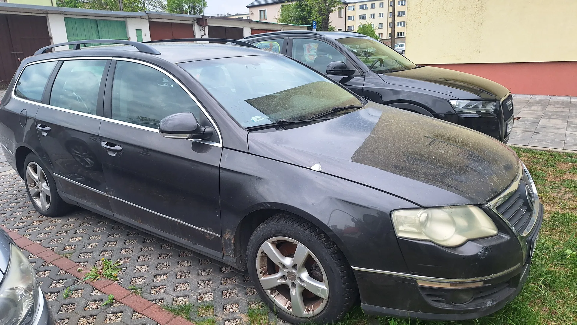 Ciemnoszary samochód typu kombi zaparkowany na podwórku z kostki brukowej. Pojazd jest zabrudzony, z widocznymi plamami na masku i bryle. W tle czarny samochód osobowy, budynki mieszkalne z zielonymi i brązowymi bramami garażowymi oraz żółta ściana z czerwoną listwą. Na pierwszym planie fragment innego pojazdu po lewej stronie. Trawa rośnie między elementami brukowymi.