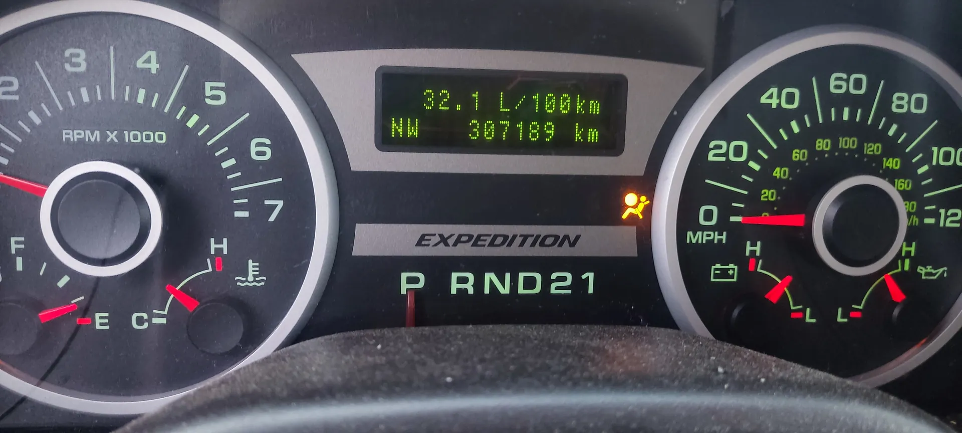 Zbliżenie na deski rozdzielcze samochodu marki Expedition. Po lewej stronie wskaźnik obrotomierza (RPM X 1000) z czerwonymi wskazówkami, oznaczeniami od 0 do 7 oraz wskaźnikiem paliwa (E-C) i temperatury (H-L). W centrum wyświetlacz cyfrowy z zielonym tekstem: "32.1 L/100km", "NW", "307189 km". Poniżej napis "EXPEDITION" oraz zielone oznaczenie biegów "P RND21". Po prawej stronie wskaźnik prędkości w MPH (0-120) z czerwoną wskazówką, dodatkowymi skalami w km/h (0-160) oraz ikonami stanu baterii i poziomu paliwa. Widoczny również żółty symbol ostrzegawczy pasów bezpieczeństwa.