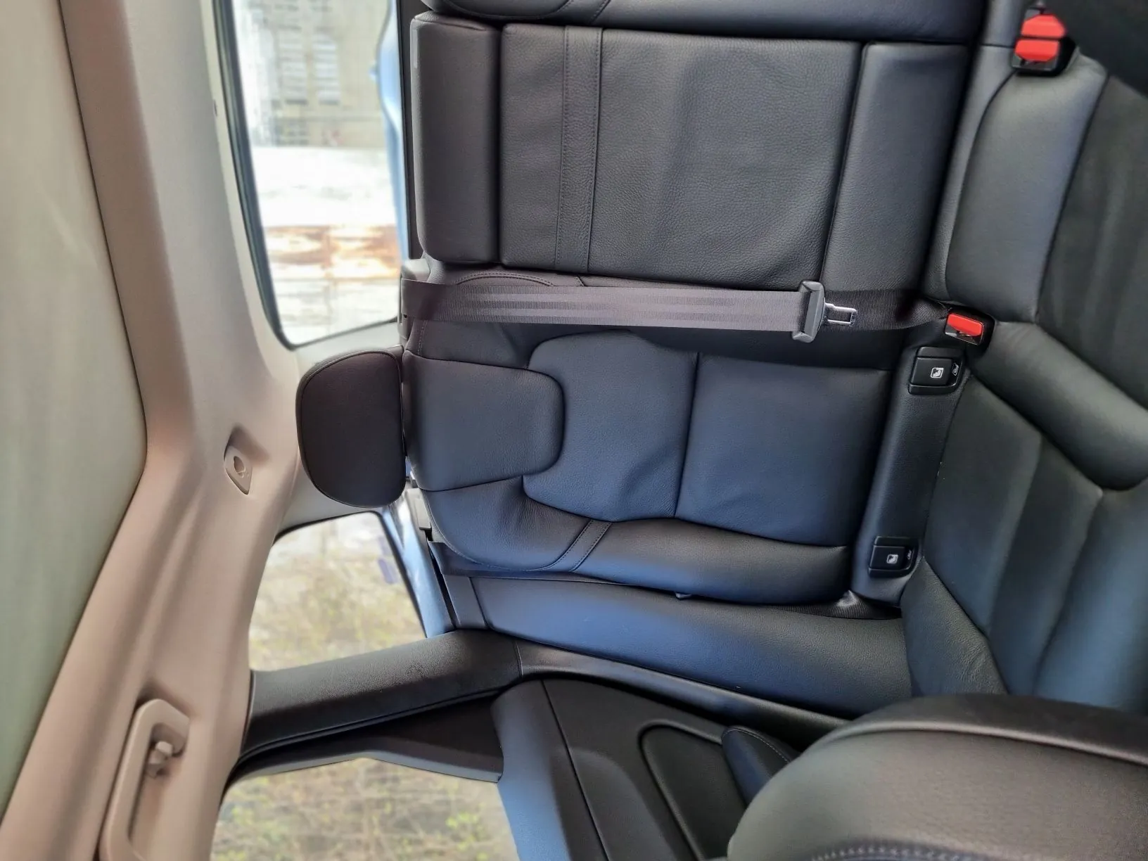 Wnętrze samochodu z widokiem na czarne skórzane siedzenia tylnego rzędu. Widoczny pas bezpieczeństwa przypięty do siedzenia, czerwone klamry zapięcia oraz fragment otwartego drzwi z beżową obudową. Przez okno widać zewnętrzne elementy: budynki i teren z zielenią. Siedzenia są ustawione pionowo, a horyzont za oknem jest poziomy.