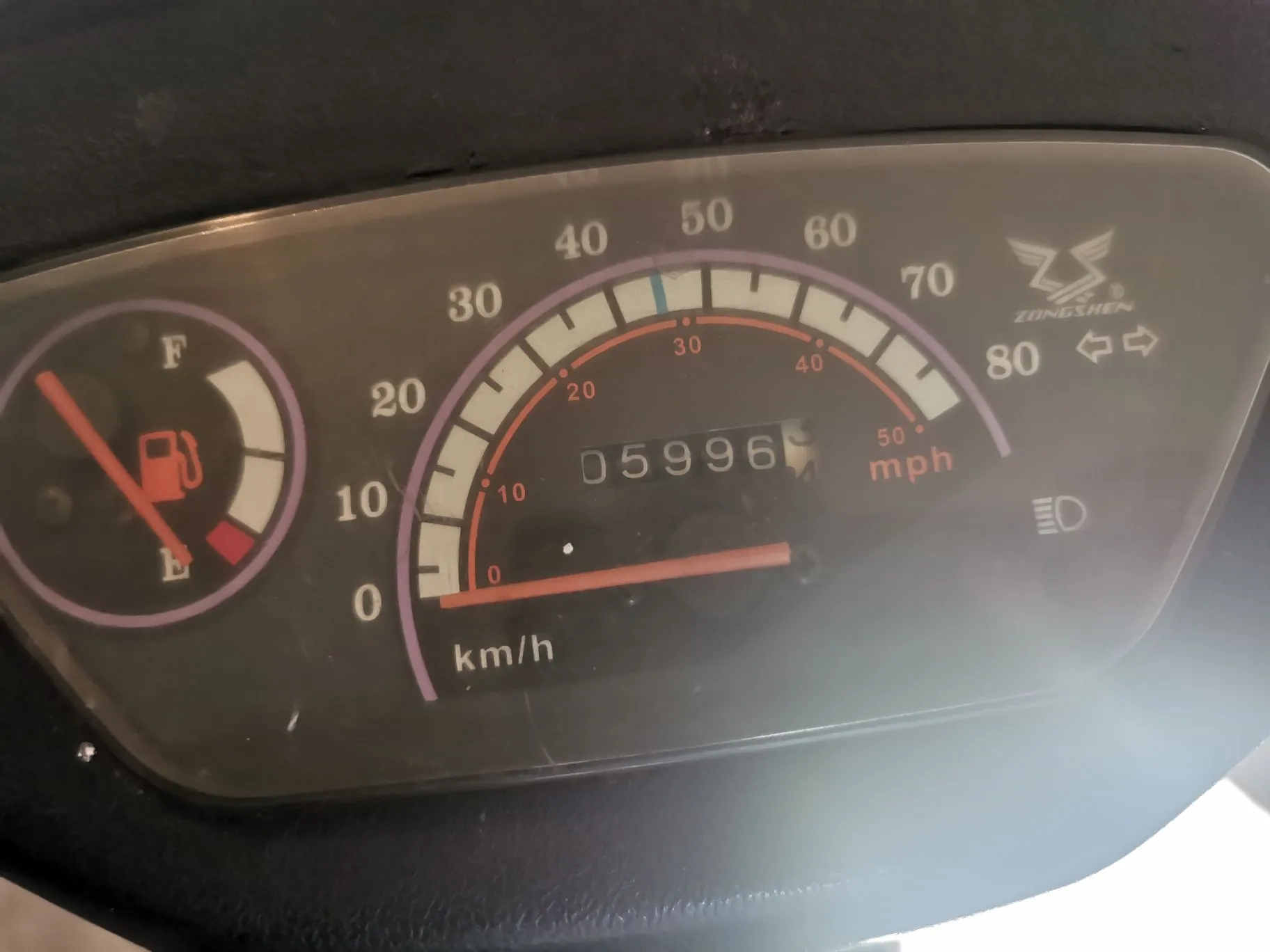 Zbliżenie na deskę rozdzielczą motocykla marki Zongshen. Po lewej stronie znajduje się wskaźnik poziomu paliwa z oznaczeniami 'F' (full) i 'E' (empty), wskazówka wskazuje poziom bliski rezerwy. W centrum znajduje się prędkościomierz z dwiema skalami: zewnętrzną w km/h (od 0 do 80) i wewnętrzną w mph (od 0 do 50). Wewnątrz prędkościomierza widoczny jest licznik przebiegu z wartością 05996. Po prawej stronie znajduje się logo Zongshen oraz ikony kierunkowskazów i świateł.