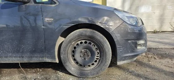 Czarny samochód osobowy z widocznym uszkodzeniem na przednim lewym błotniku. Samochód jest zabrudzony, z czarną stalową felgą i oponą z widocznymi śladami zużycia. Tło stanowi betonowa ściana i brudna nawierzchnia ziemi. Widoczne są reflektory oraz fragment przedniej maski.