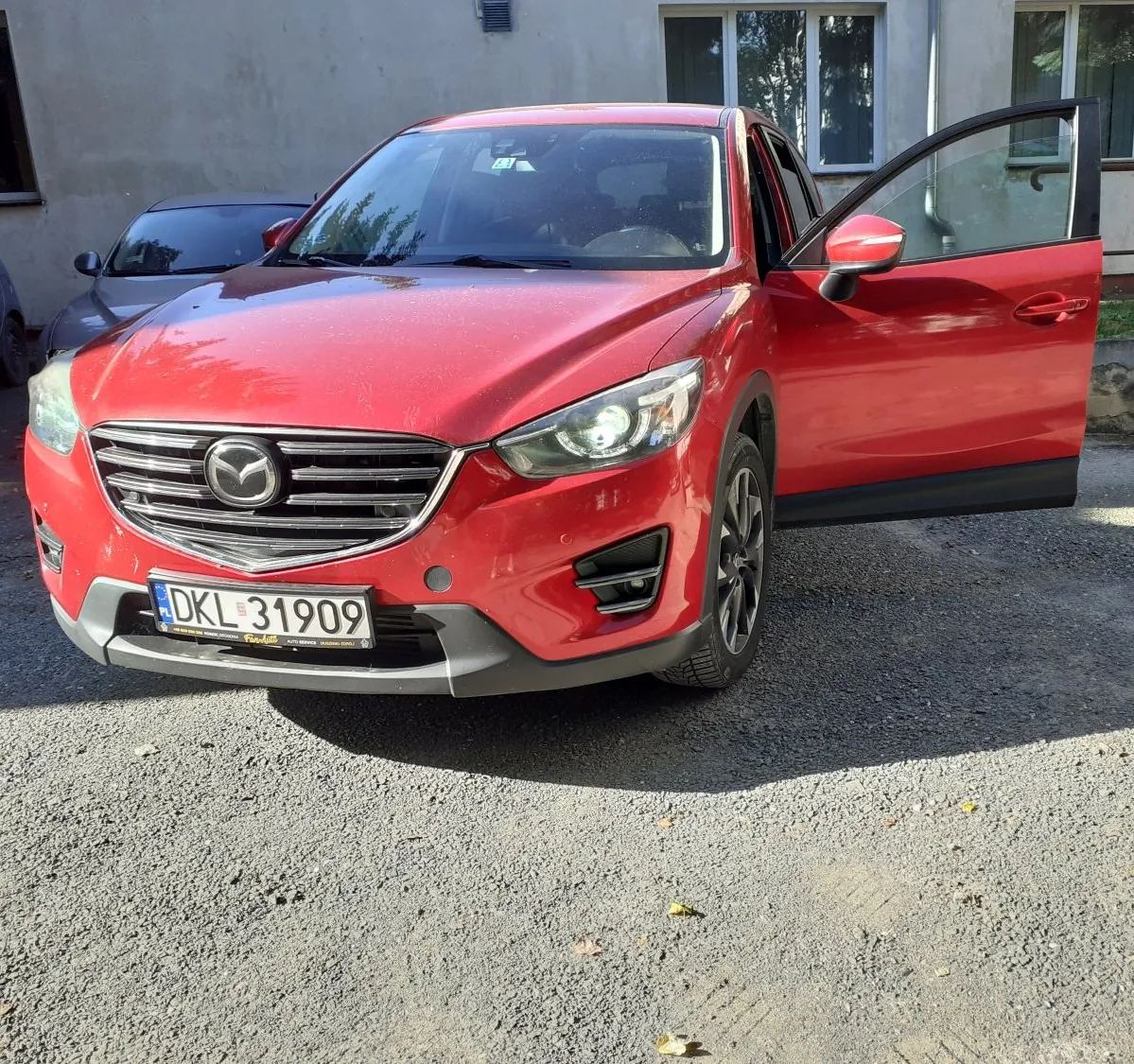 Czerwony samochód marki Mazda zaparkowany na żwirowym podjeździe przed jasnym budynkiem. Przednie prawe drzwi auta są otwarte. Samochód posiada tablicę rejestracyjną o numerze DKL 31909. W tle widoczny jest fragment innego, ciemnego samochodu oraz okna budynku.
