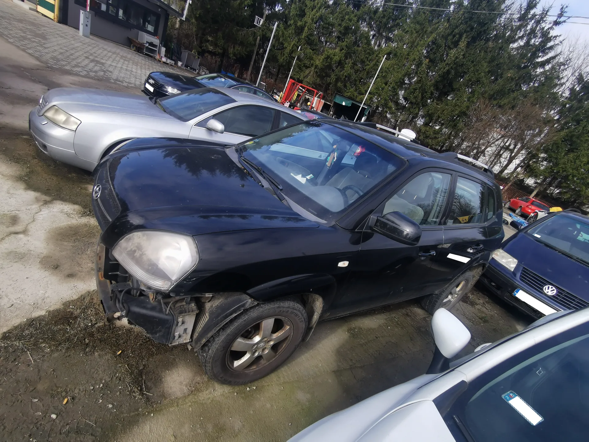 Parking z kilkoma samochodami, w tym czarnym SUV-em z uszkodzonym przodem (uszkodzony zderzak i reflektor). W tle srebrny sedan, ciemnoniebieski samochód marki Volkswagen oraz inne pojazdy. Tło stanowią drzewa, budynek i sprzęt. Podłoga parkingowa składa się z betonu i ziemi.