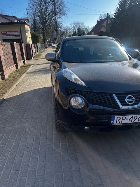 Czarny samochód marki Nissan zaparkowany na brukowanej ulicy. Widoczna tablica rejestracyjna z napisem RP 49. W tle budynki, drzewa bez liści i jasne niebo. Po lewej stronie ulicy znajduje się chodnik oraz ogrodzenie z cegły.