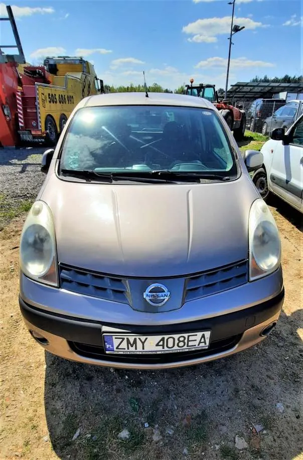 Przedni widok srebrnego samochodu marki Nissan z tablicą rejestracyjną ZMY 408EA. W tle widoczne są inne pojazdy: żółty ciężarówka z numerem 505 606 997, czerwony traktor oraz biały samochód. Scena rozgrywa się na otwartym terenie z żwirem i trawą pod błękitnym niebem z chmurami.