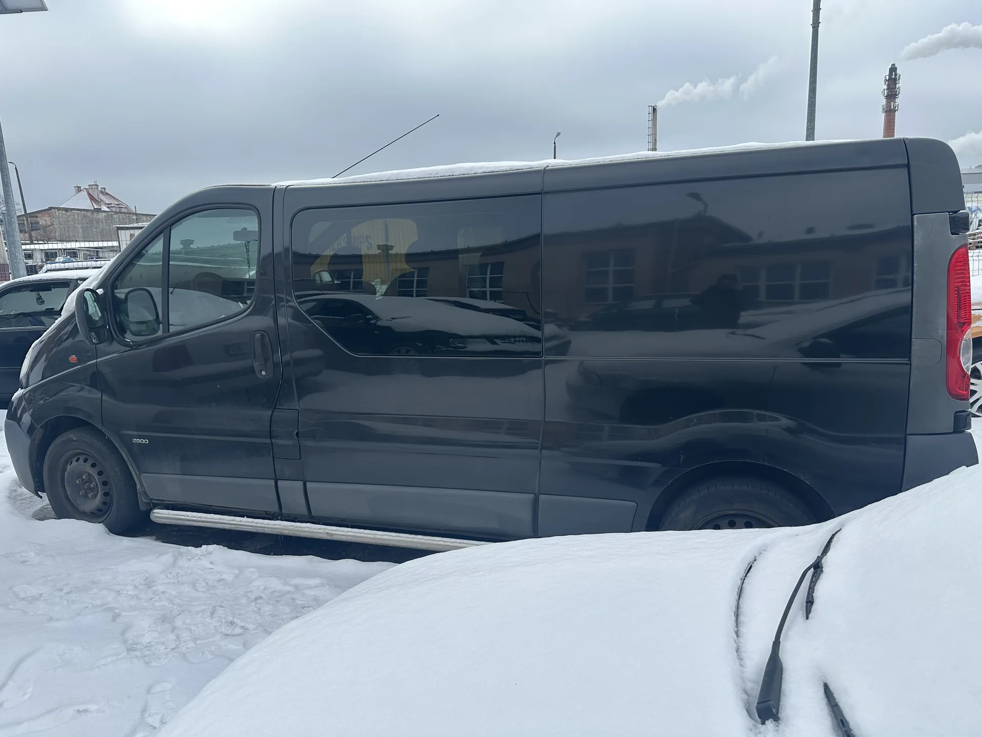 Ciemnoszary furgon marki z widoczną cyfrą 2500 na przednim błotniku, zaparkowany na śnieżnej powierzchni. W tle budynki z dachami pokrytymi śniegiem, przemysłowe kominy z dymem oraz inne pojazdy zakryte śniegiem. Niebo jest zachmurzone. Na boku furgona widoczne są odbicia budynków i innych samochodów.