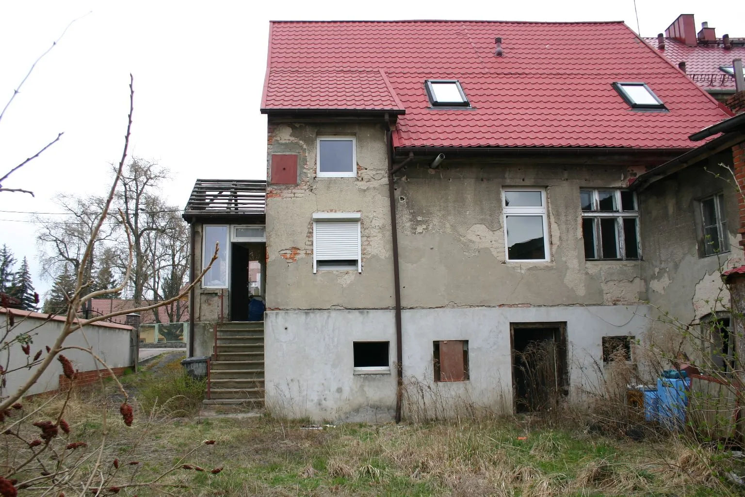 Zdjęcie starego, dwukondygnacyjnego budynku z czerwonym, spadzistym dachem z dwoma oknami dachowymi. Elewacja budynku jest w złym stanie, z widocznymi ubytkami tynku i odsłoniętą cegłą. Na parterze znajdują się ciemne otwory drzwiowe i okienne, a na piętrze kilka okien, w tym jedno z białą roletą zewnętrzną. Po lewej stronie widoczne są betonowe schody prowadzące do wejścia. Przed budynkiem znajduje się nieuporządkowany teren z suchą trawą i krzewami.