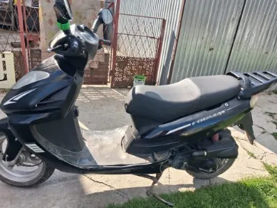 Czarny skuter zaparkowany na betonowej powierzchni, z widoczną trawą w dolnej części kadru. W tle metalowe ogrodzenie i brama z siatką. Skuter ma czarne siedzenie, przednią maskę oraz widoczne elementy konstrukcyjne. Na boku skutera znajduje się nieczytelny napis.