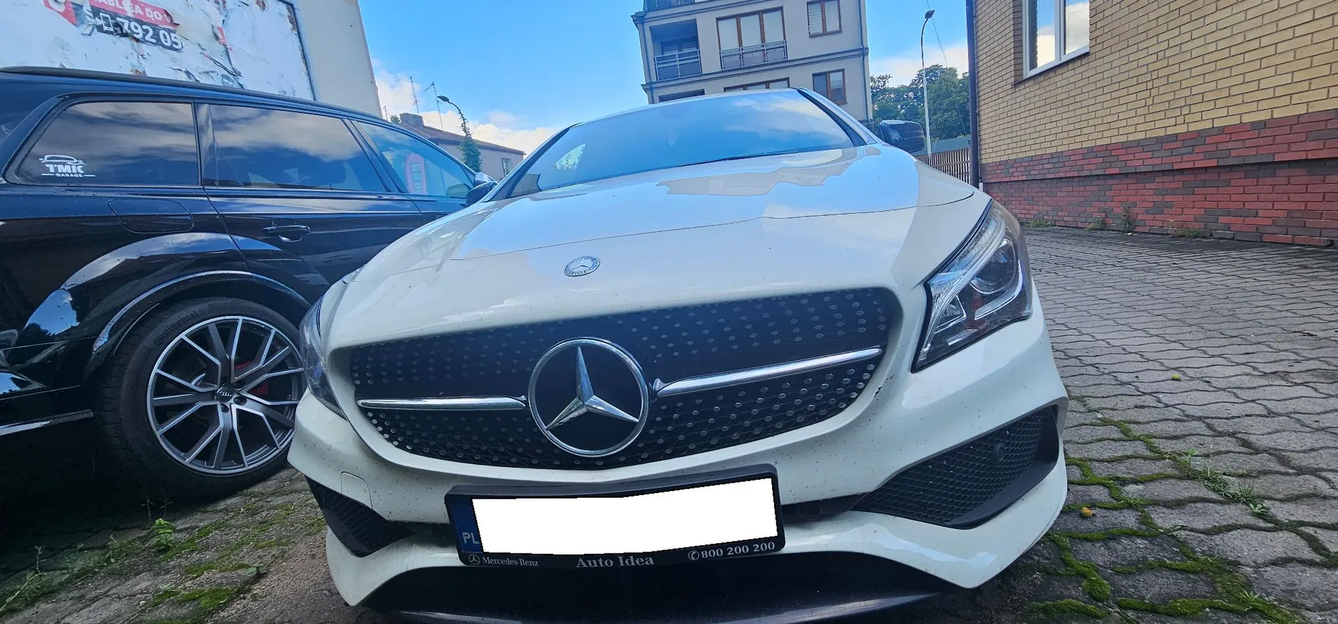 Zdjęcie przodu białego samochodu marki Mercedes-Benz zaparkowanego na kostce brukowej. W centralnej części grilla znajduje się duże logo marki. Po lewej stronie widoczny jest fragment czarnego samochodu. W tle znajduje się jasny budynek mieszkalny oraz fragment ściany z cegieł. Na dolnej ramce tablicy rejestracyjnej widnieje napis 'Auto Idea' oraz numer telefonu '800 200 200'.