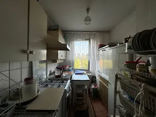 Kuchnia z białymi szafkami wiszącymi po lewej stronie, kuchenką gazową z czajnikiem, zlewozmywakiem i deska do krojenia na blacie. W centrum stoi stół z krzesłem, za nim okno z białymi zasłonami wpuszczającymi światło. Po prawej stronie widoczna lodówka oraz metalowe półki z naczyniami i kuchenkami. Podłoga wyłożona kafelkami, sufit z wiszącą lampą. Całość w neutralnych barwach, bez widocznego tekstu.