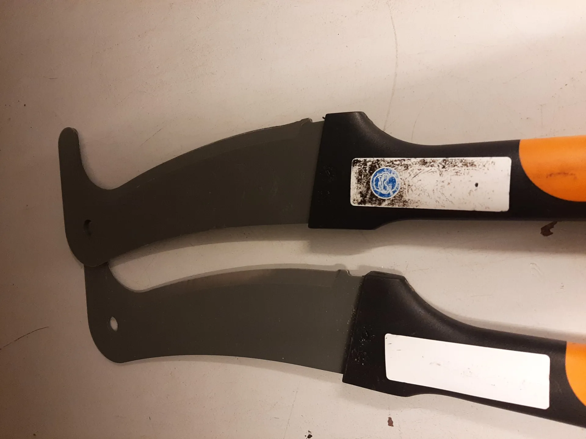 Dwa noże ogrodnicze z czarnymi ostrzami i czarno-pomarańczowymi uchwytami leżące na jasnej powierzchni. Każdy nóż ma białą etykietę na uchwycie; na prawym nożu etykieta jest zabrudzona, a na niej widoczny niebieski okrągły znak z białym symbolem. Ostrza mają zakrzywiony kształt z otworem blisko uchwytu.