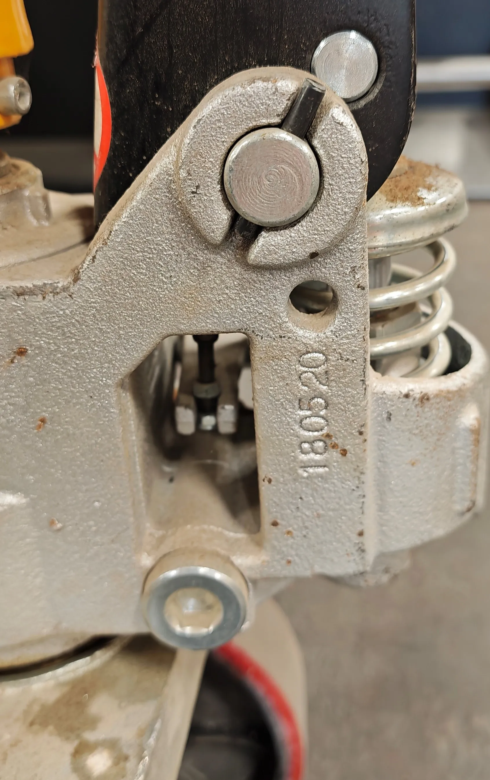 Zbliżenie na metalowy element mechaniczny w kolorze srebrnym. Widoczny jest zawias z bolcem, sprężyna oraz wybity numer seryjny 180520. Element jest połączony z czarną belką.