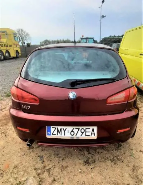 Zdjęcie przedstawia tył czerwonego samochodu marki Alfa Romeo 147 z tablicą rejestracyjną ZMY 679EA. Samochód stoi na brudnej, piaszczystej powierzchni. W tle widoczne są dwa żółte pojazdy (pojazd ciężarowy po lewej i furgon po prawej), ogrodzenie, drzewa oraz słup z latarnią. Niebo jest pochmurne. Na tylnym szybie samochodu widoczne są ślady zabrudzeń.
