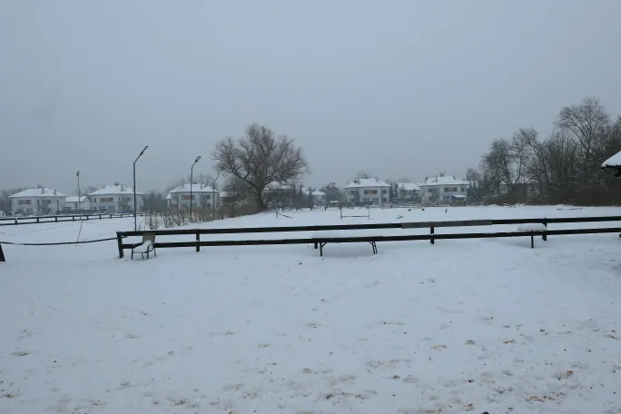 Zimowy, zamglony krajobraz z pokrytym śniegiem terenem. Na pierwszym planie znajduje się zaśnieżona powierzchnia z widocznymi śladami stóp, oddzielona niskim, ciemnym drewnianym ogrodzeniem. Za ogrodzeniem widać kilka białych, nowoczesnych domów jednorodzinnych z dwuspadowymi dachami oraz bezlistne drzewa. Niebo jest jednolicie szare i zamglone.