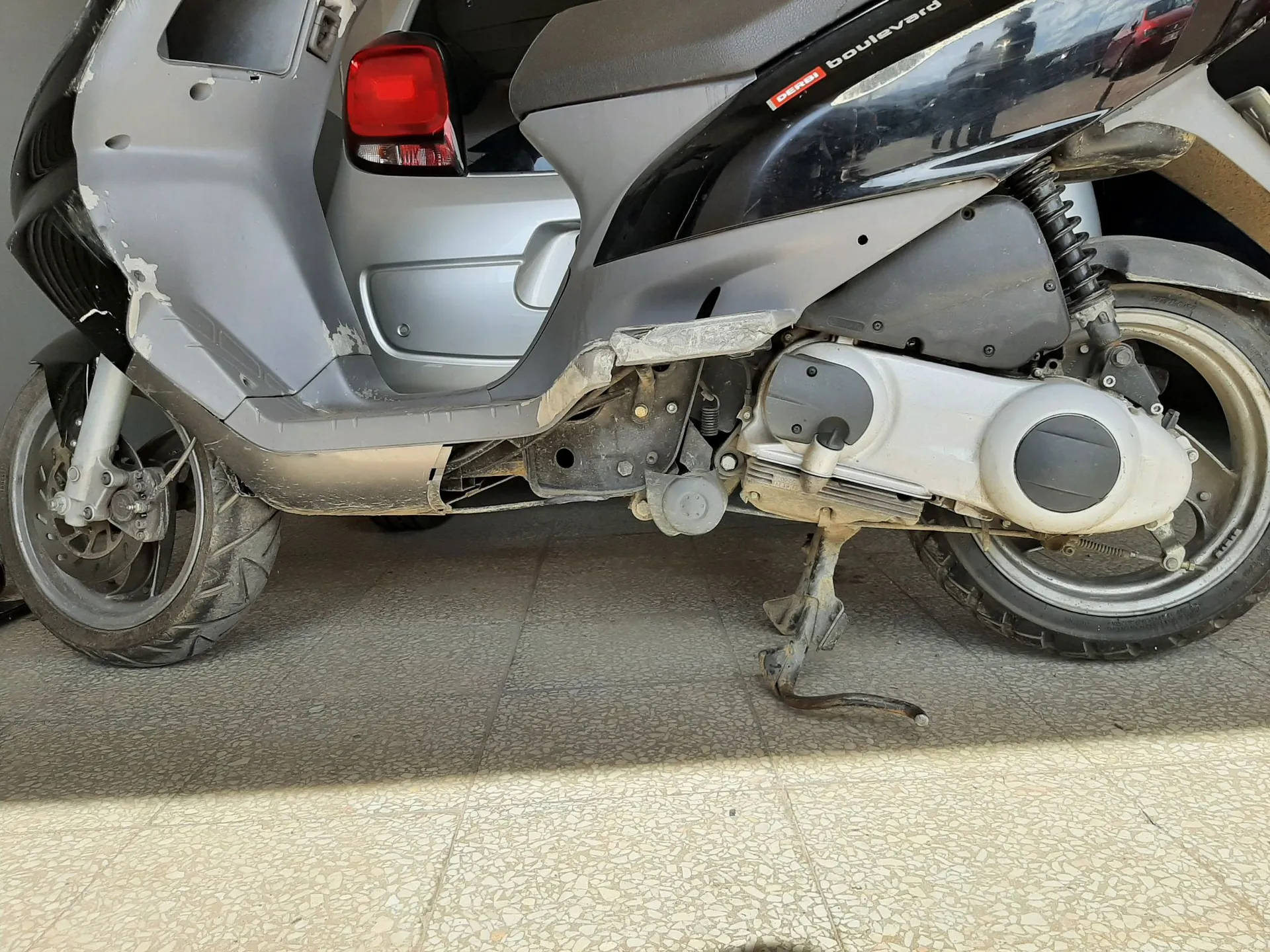 Zbliżenie na prawy bok srebrno-czarnego skutera marki Derbi, model Boulevard. Pojazd posiada liczne uszkodzenia: pęknięcia i ubytki w srebrnych plastikach owiewki z przodu oraz w dolnej części nadwozia. Skuter stoi na bocznej stopce na jasnej, kafelkowej powierzchni.