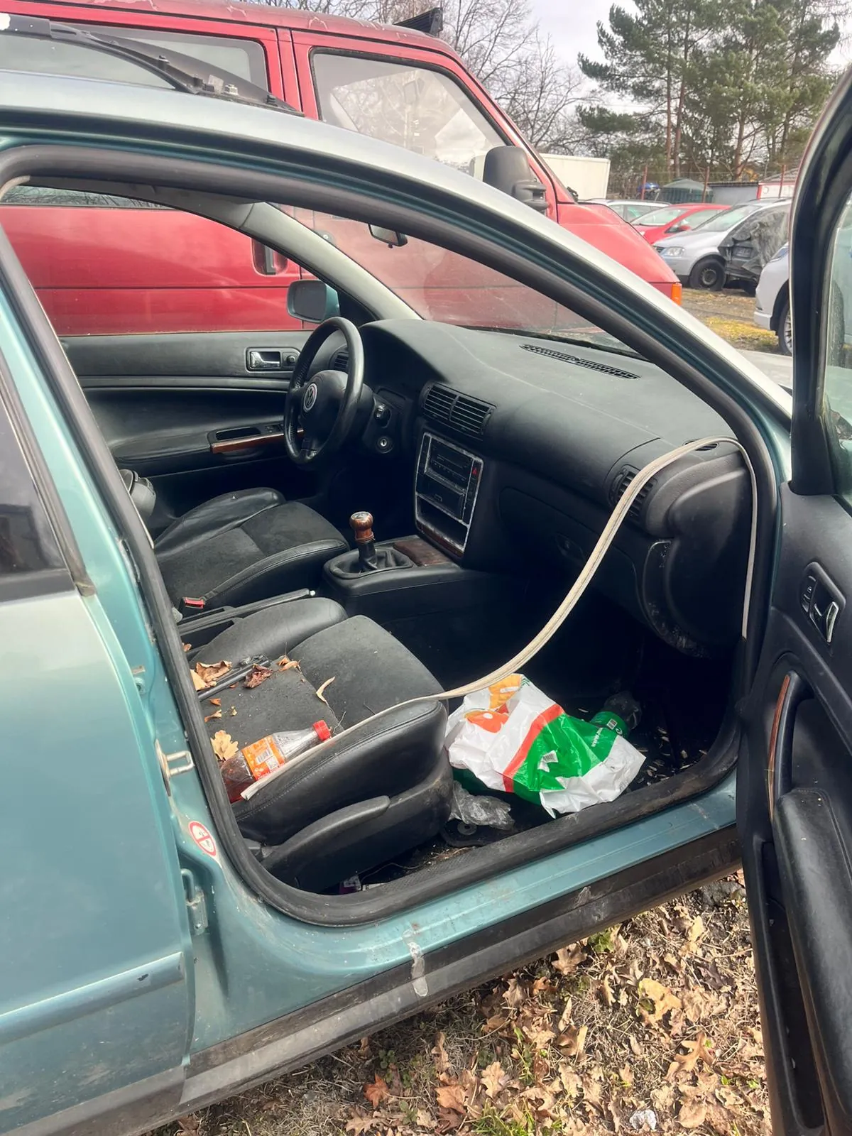 Wnętrze zielonego samochodu z otwartymi drzwiami. Widoczne czarne siedzenia, kierownica, deska rozdzielcza z radioodtwarzaczem i dźwignią zmiany biegów z drewnianą rączką. Na przednim siedzeniu pasażera leży plastikowa butelka z czerwoną nakrętką oraz suchy liść. Na podłodze samochodu znajduje się torba zielono-biała z pomarańczowym elementem. W tle czerwony van, inne zaparkowane samochody, drzewa i sucha trawa na ziemi.