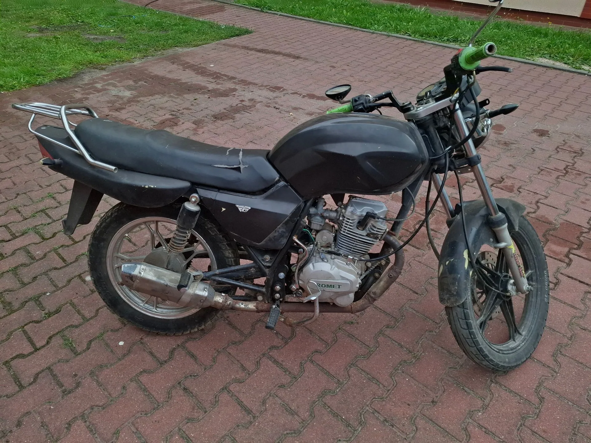 Czarny motocykl marki Romet zaparkowany na kostce brukowej. Pojazd posiada czarny bak, siedzenie oraz błotniki. Widoczny jest srebrny wydech, silnik z napisem ROMET oraz metalowy bagażnik z tyłu. Kierownica ma zielone gumowe uchwyty.