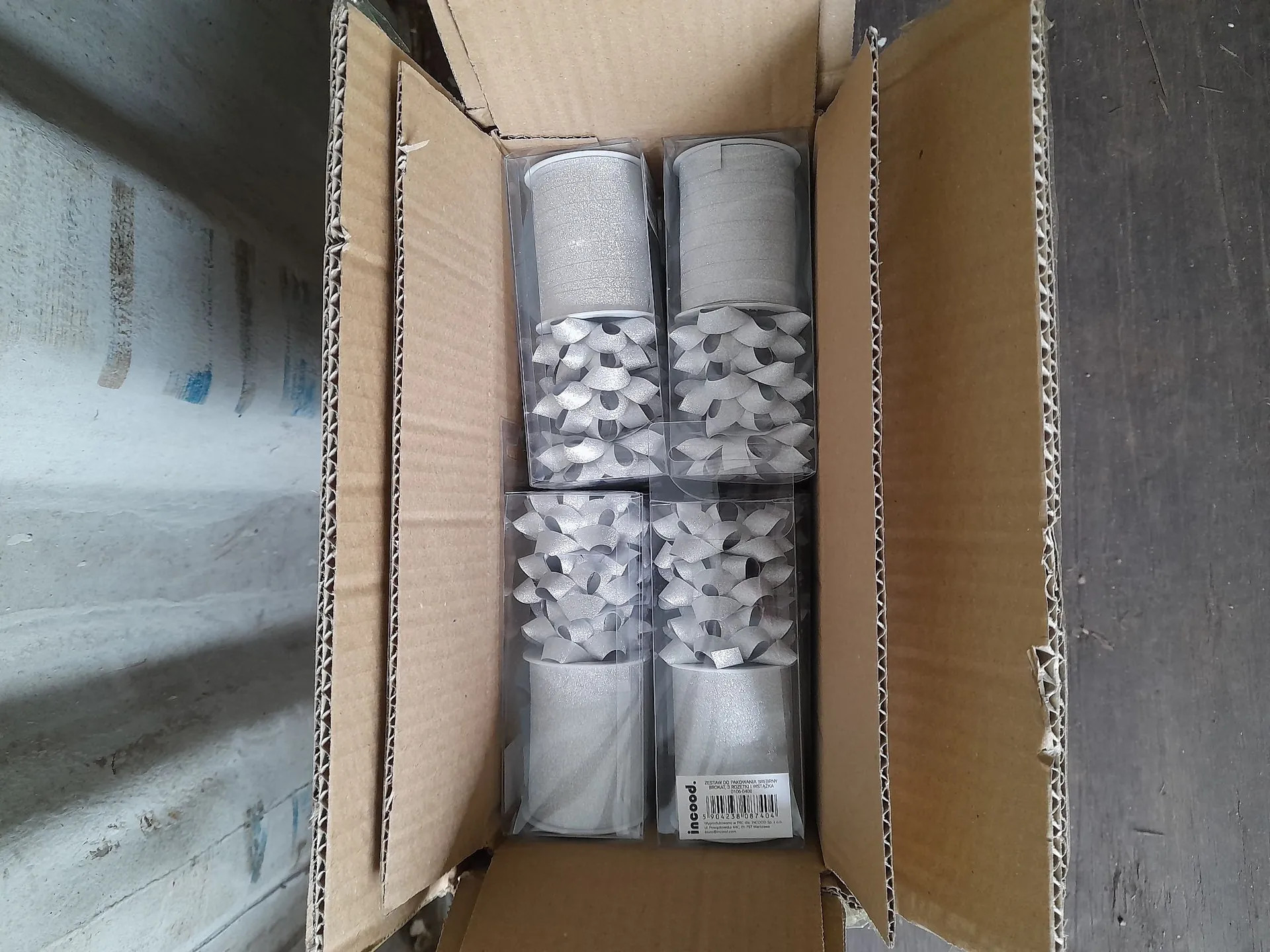 Widok z góry do wnętrza otwartego kartonu z tektury, w którym znajdują się cztery przezroczyste plastikowe opakowania. W każdym opakowaniu znajduje się biały, cylindryczny przedmiot z ozdobnymi, spiralnymi wycięciami w środkowej części. Na jednym z opakowań widoczna jest biała etykieta z kodem kreskowym i napisem 'inwood'.